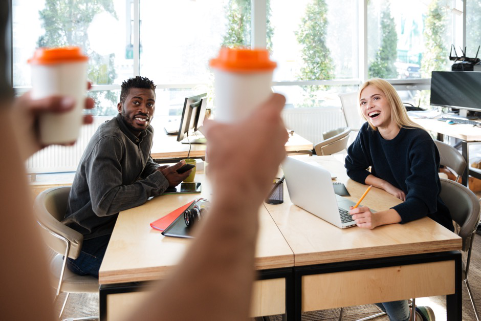 Two employees smile as a coworker brings them coffee at their desk.