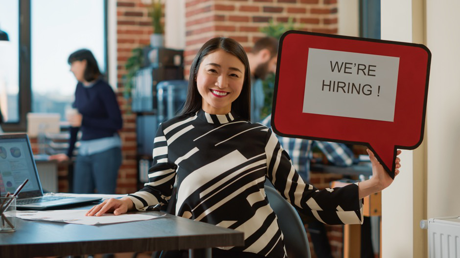  A woman smiles, holding a sign that reads "we're hiring!"