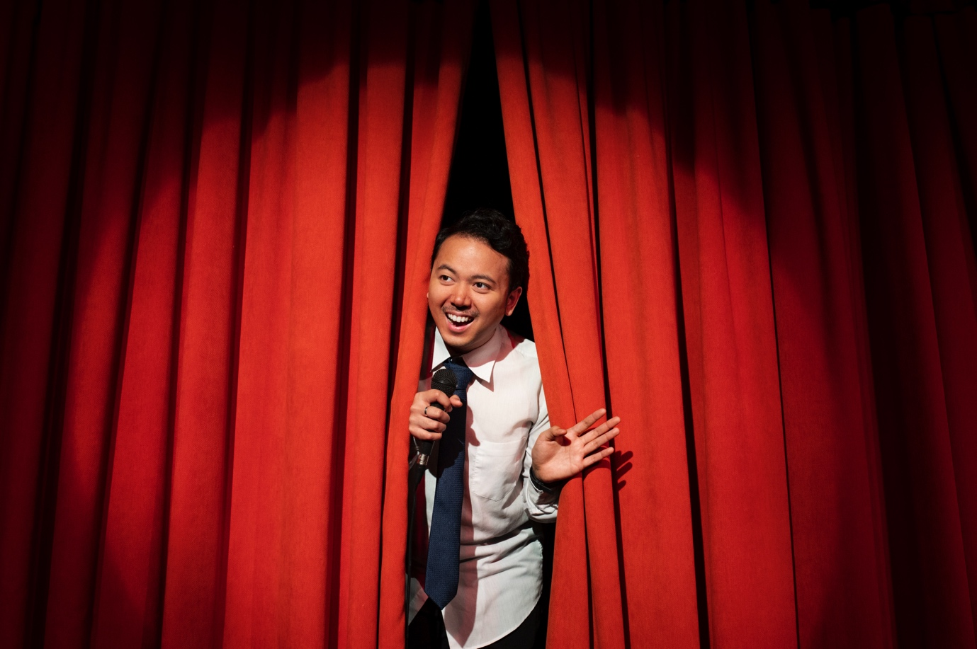 A stand-up comedian peers through a red curtain, about to go onstage.