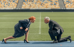 A businesswoman and a businessman face off on a track with a soccer field in the background.