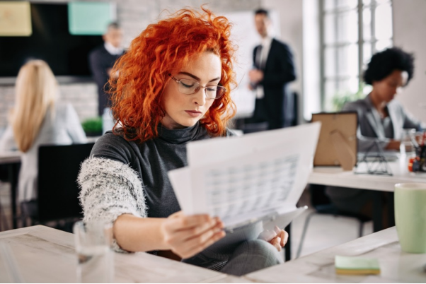 a woman with orange hair analyzes data on paper and on her computer